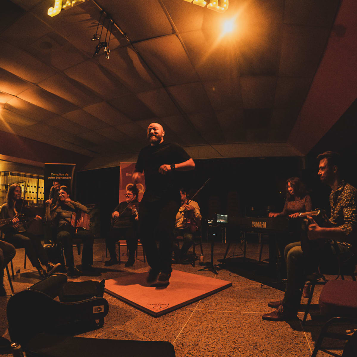 Festival de musique Trad Val-d'Or - Un homme danse sur une planche de bois, entouré de musiciens.