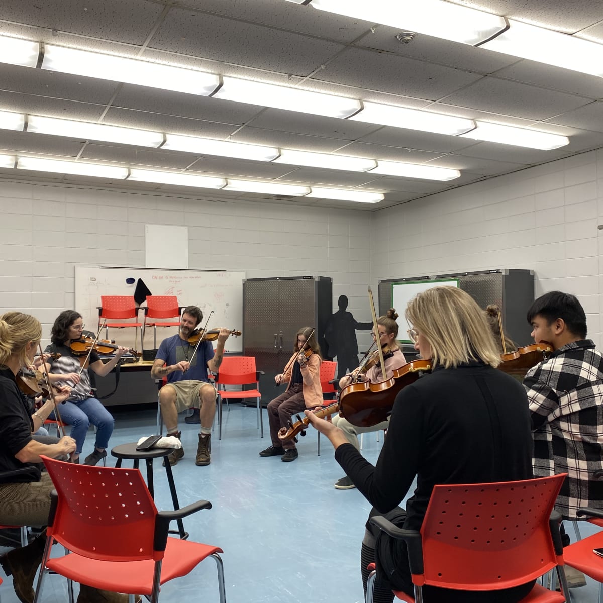 Festival de musique Trad Val-d'Or - Des individus sont assis en cercle sur des chaises rouges et jouent du violon.