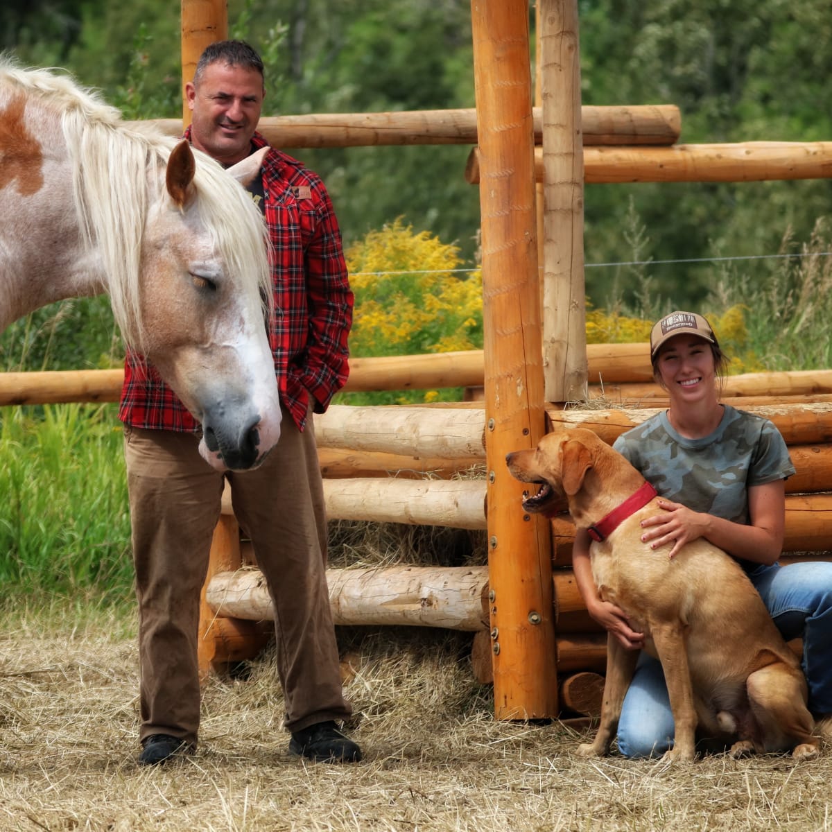 A man stands next to a horse, and next to him, a woman crouches with a dog.