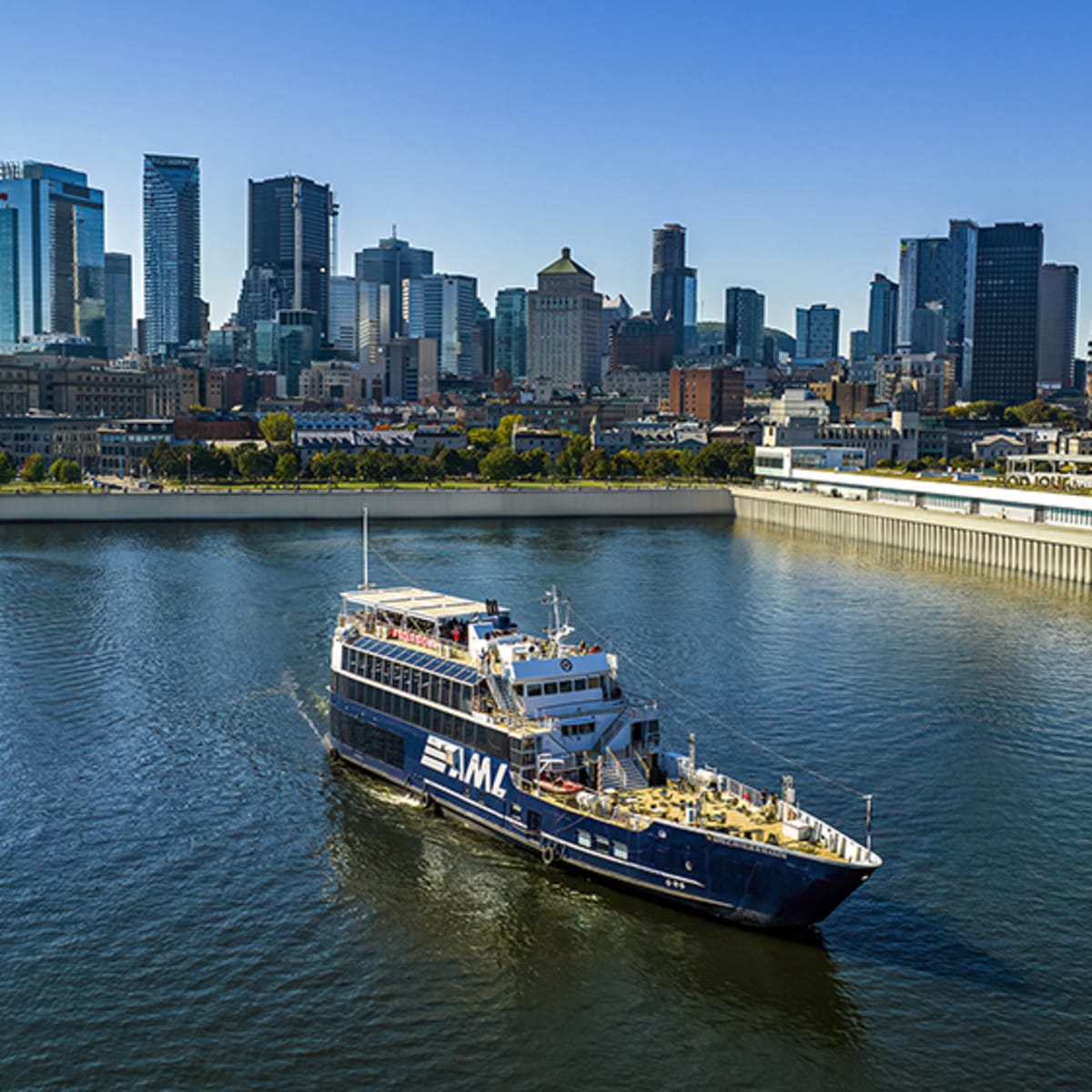 Un bateau de croisière naviguant sur une rivière, avec une vue panoramique sur une ville avec de hauts gratte-ciels en arrière-plan sous un ciel dégagé.