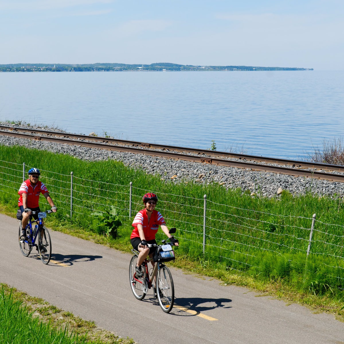 Two cyclists on a paved path bordered by a railroad track and a waterway.