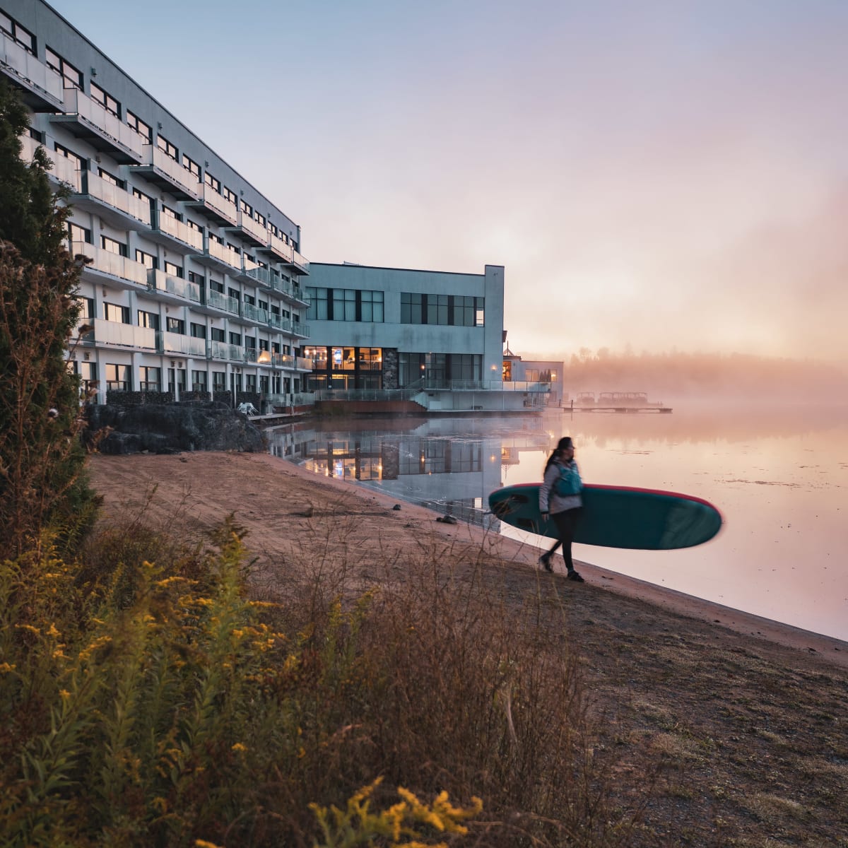 A woman holding a paddleboard at the edge of a lake at dawn.