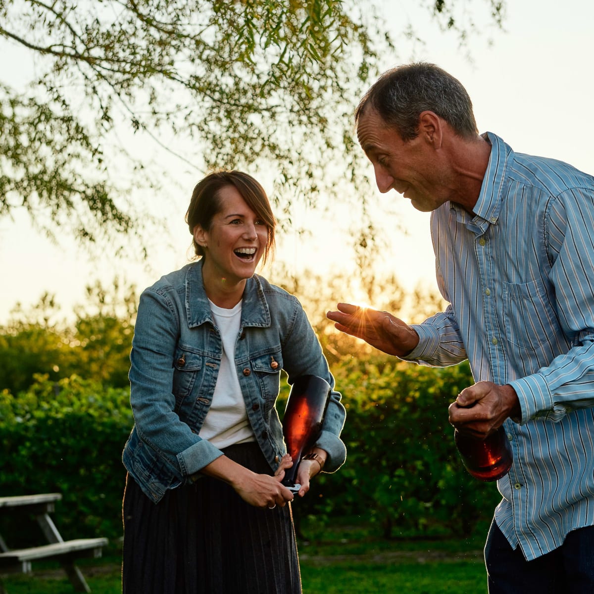 At sunset, a smiling woman and man are each holding a bottle of wine.
