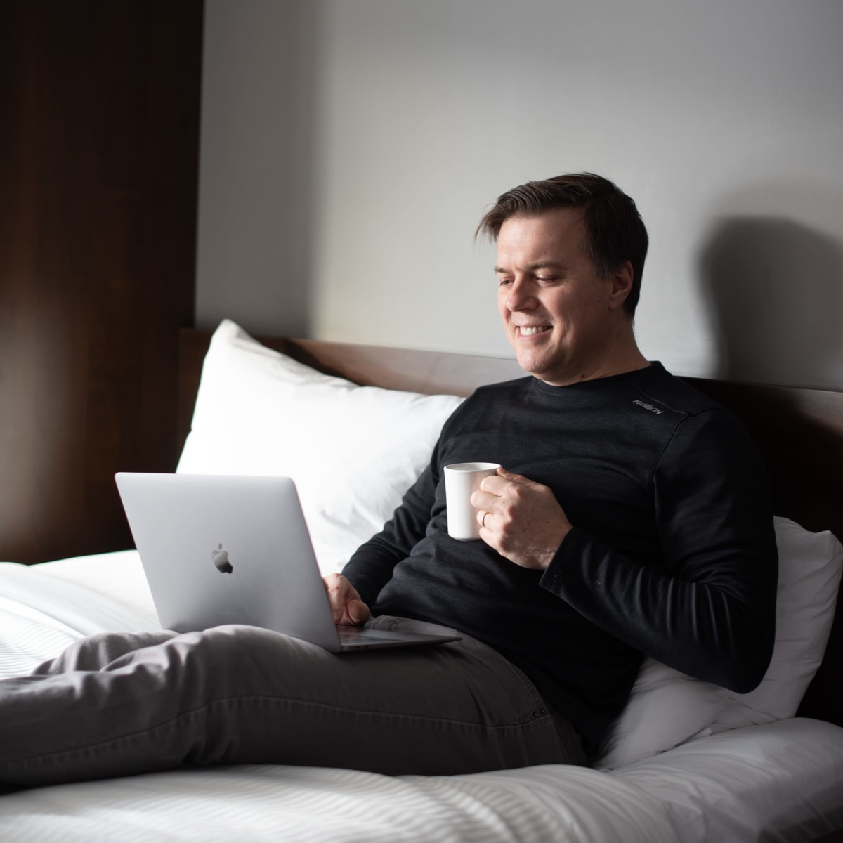 Homme allongé sur un lit, avec un café en main et un ordinateur portable sur ses cuisses.