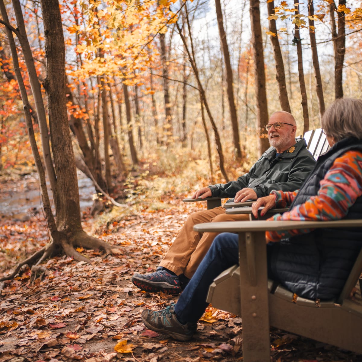 Municipalité d'Orford - A couple sitting on wooden chairs in a forest with autumn colors
