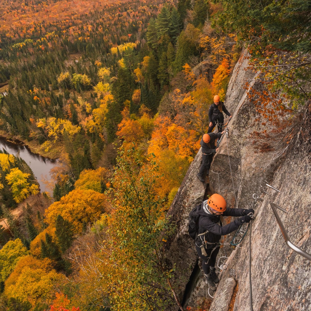 Personnes escaladant une paroi rocheuse de la Via Ferrata du Diable, avec un paysage d'automne en arrière-plan.