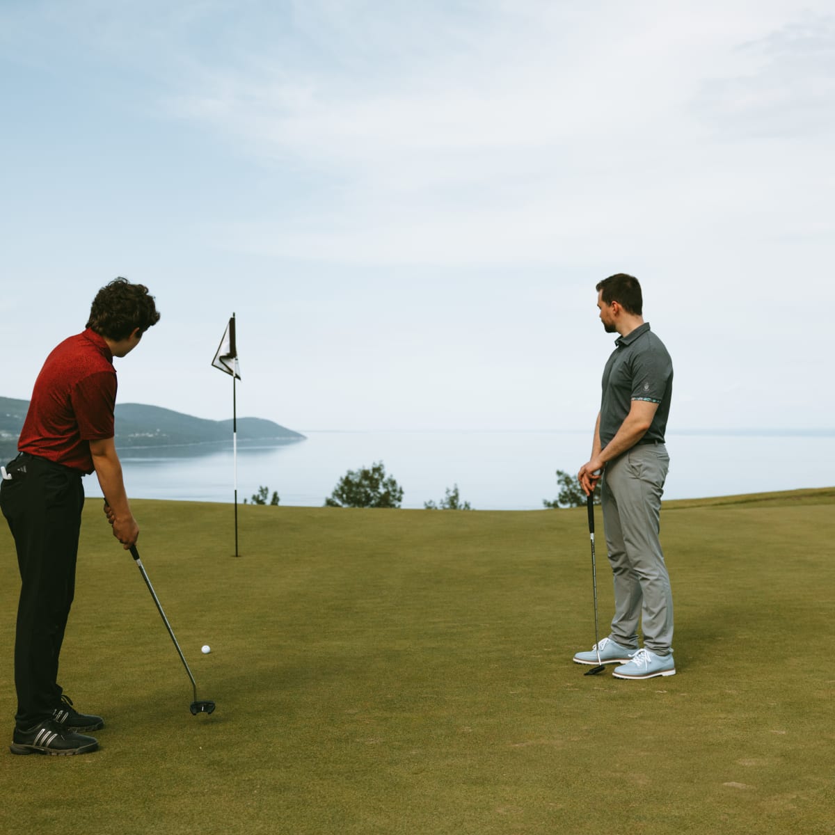 Jeune homme qui s'apprête à jouer un coup au Golf Manoir Richelieu, son père le regarde et le fleuve s'étend derrière eux.