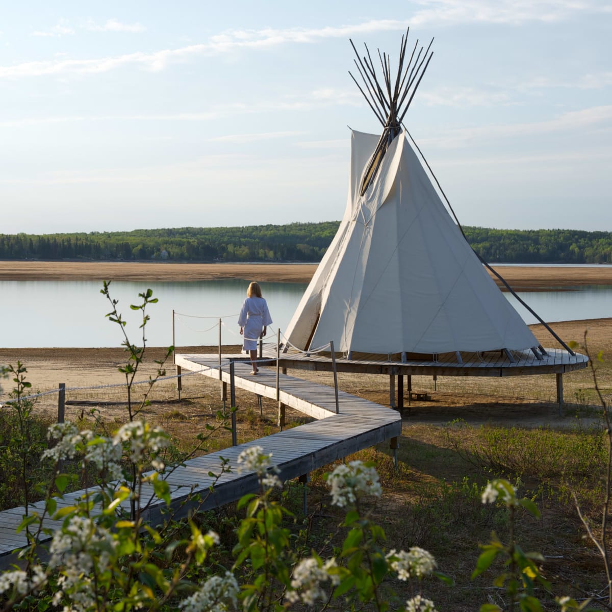 A woman in a bathrobe walking beside a teepee on the shore of a lake in the summer.  