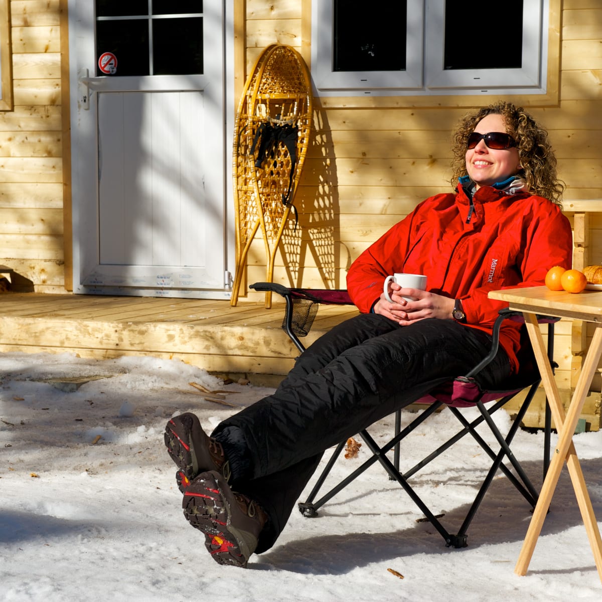 Woman sitting outside in the winter in the sun with some breakfast and snowshoes behind her..