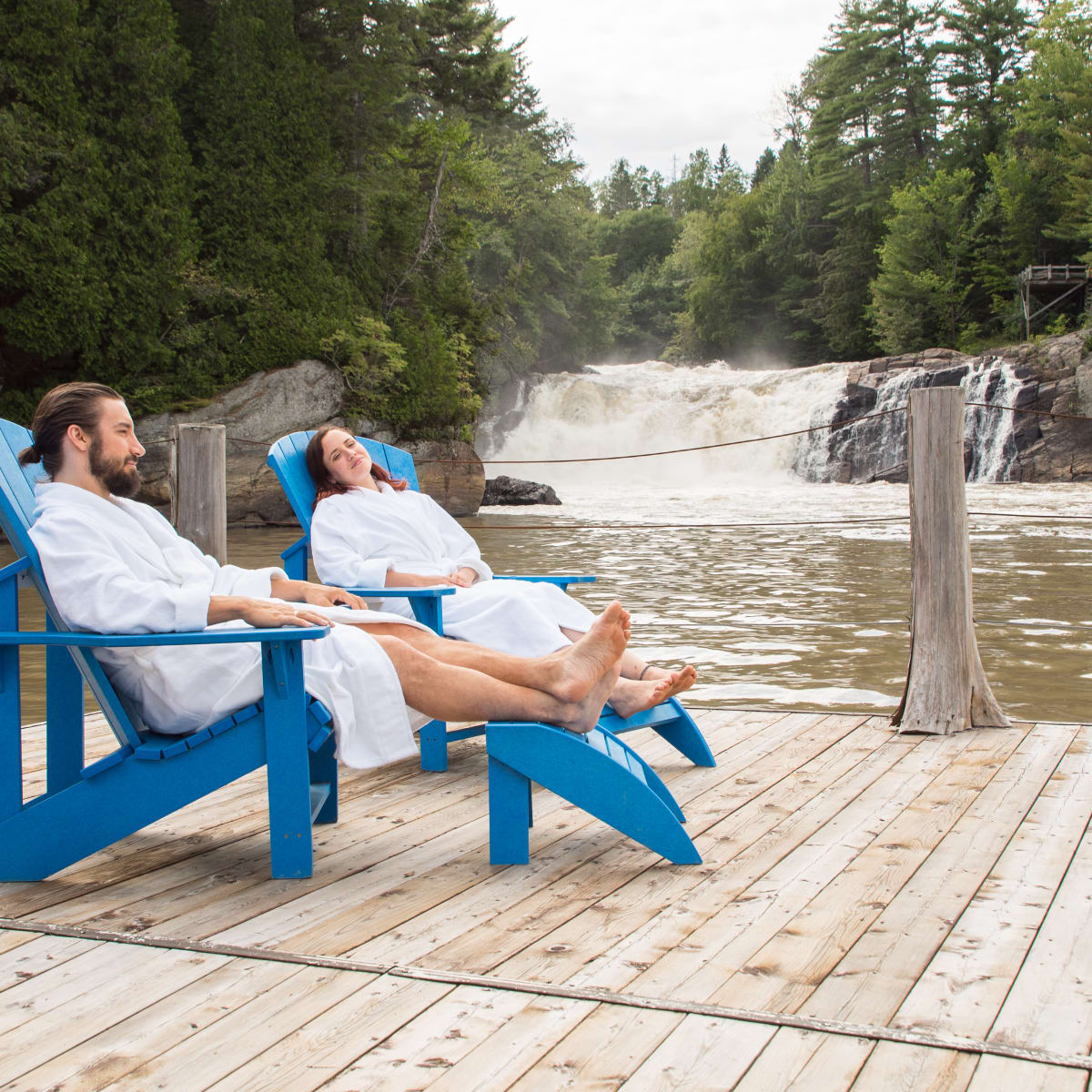 Couple in bathrobes on patio chairs on a deck next to a waterfall.