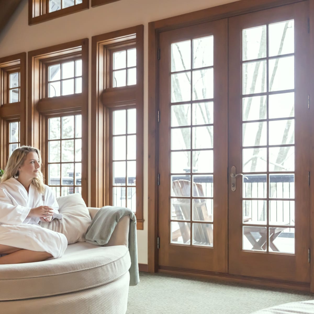 Woman in bathrobe sitting on a comfy chair in a room with many windows.