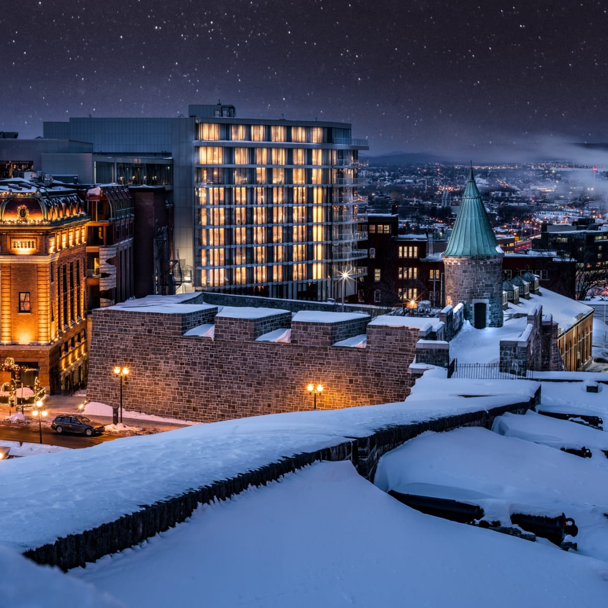 L’édifice moderne de l’hôtel parmi des bâtiments historiques sous la neige; le ciel est étoilé et la ville est illuminée au loin.