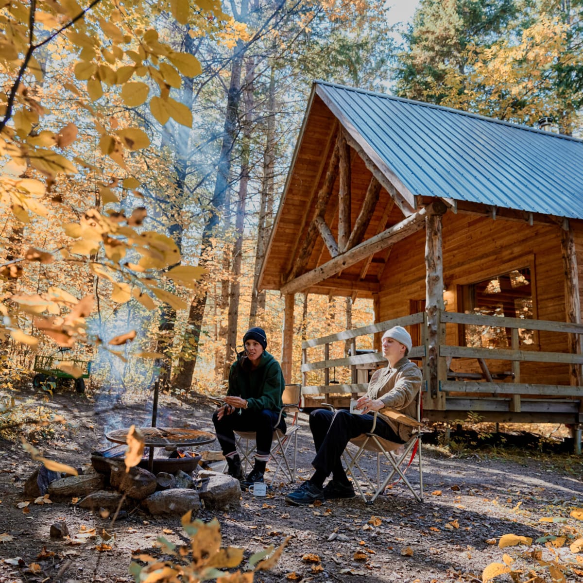 Two people sitting around a campfire in front of a wooden cottage in a forest in autumn.