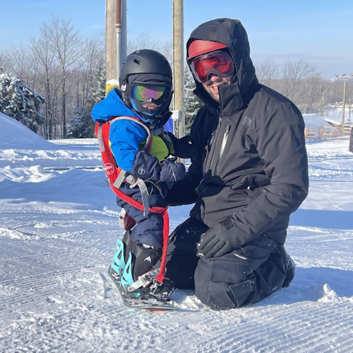 Un père avec son garçon, le bambin a une planche à neige aux pieds et le père est agenouillé devant lui.