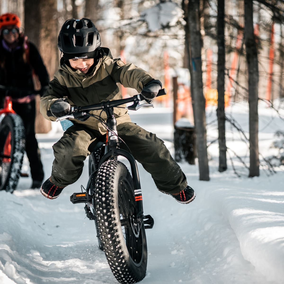 Young boy on a fatbike in a snow-covered forest in the foreground, with another person further behind.