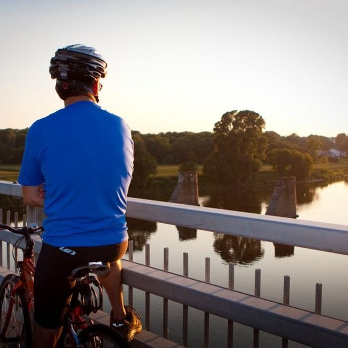 Homme à vélo vu de dos; il semble de tenir sur un pont au-dessus de l’eau.