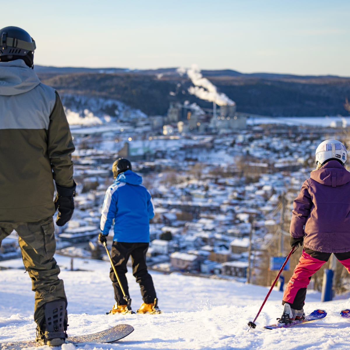 Père, fils et fille en habits de ski sur la montagne avec la vue du centre-ville devant eux.