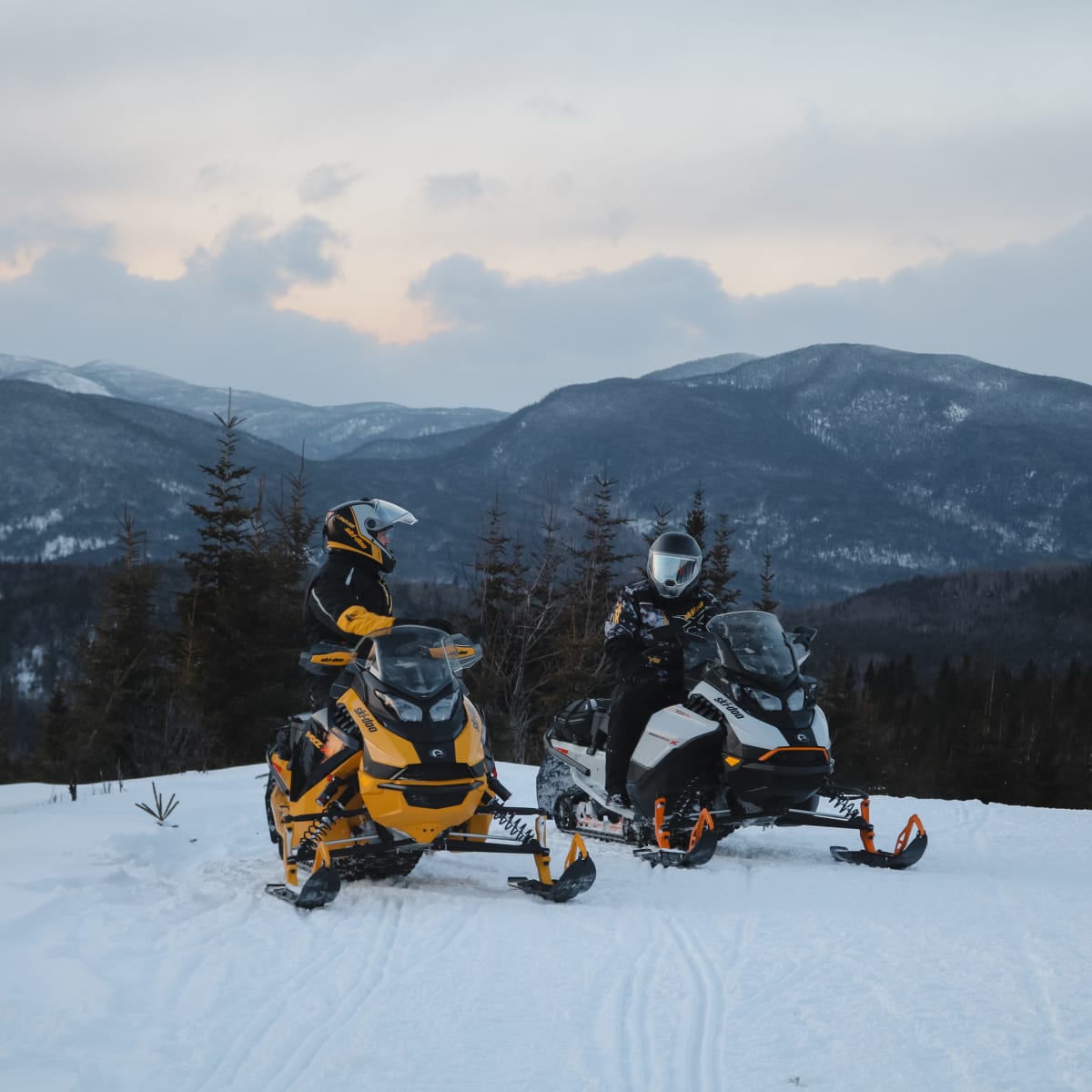 Two snowmobilers on a trail, with mountains in the background.