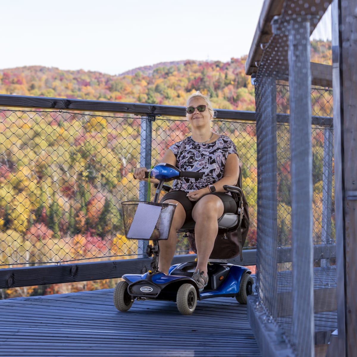 Femme en quadriporteur sur une passerelle en bois du Sentier des cimes, devant un paysage coloré.