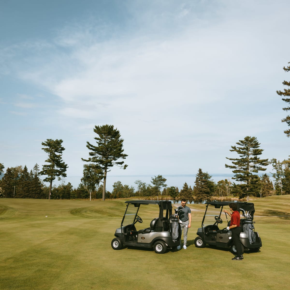 Two golf carts are parked on a golf course; two men are standing next to them.