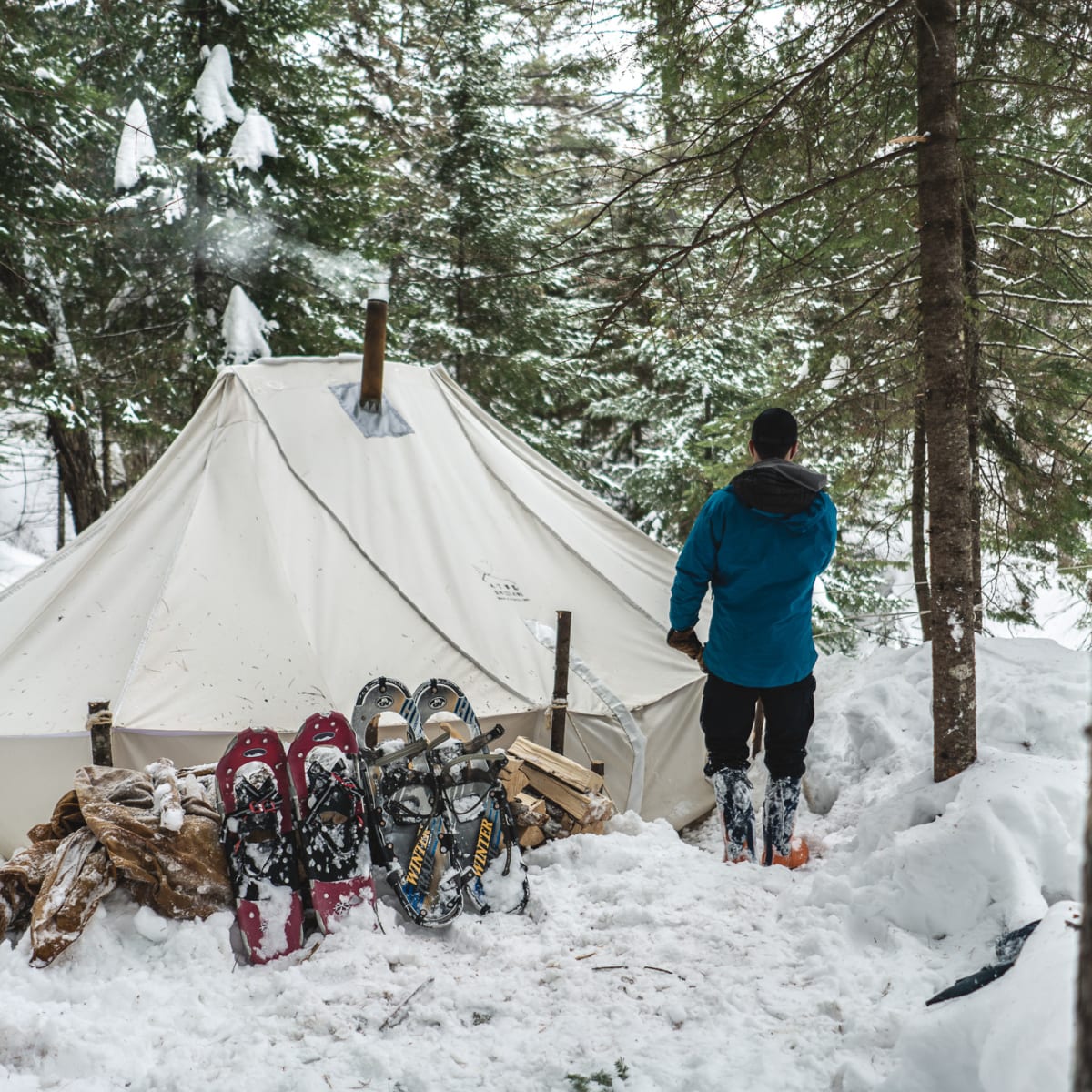 Personne devant une tente de camping hivernal, avec deux paires de raquettes plantées dans la neige à ses côtés.
