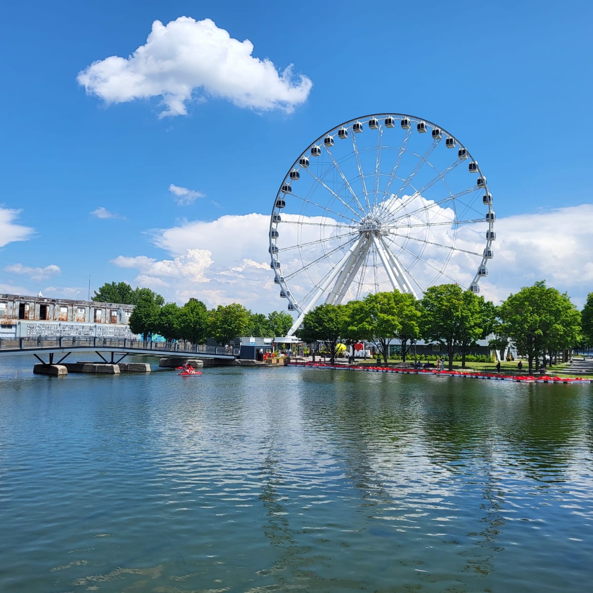 Grande Roue de Montréal vue de loin, avec un plan d'eau à l'avant et un ciel bleuté à l'arrière.