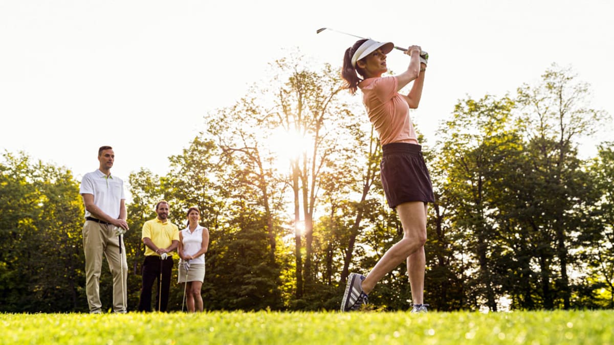 Une femme joue au golf, avec trois autres individus qui l'observent en arrière-plan