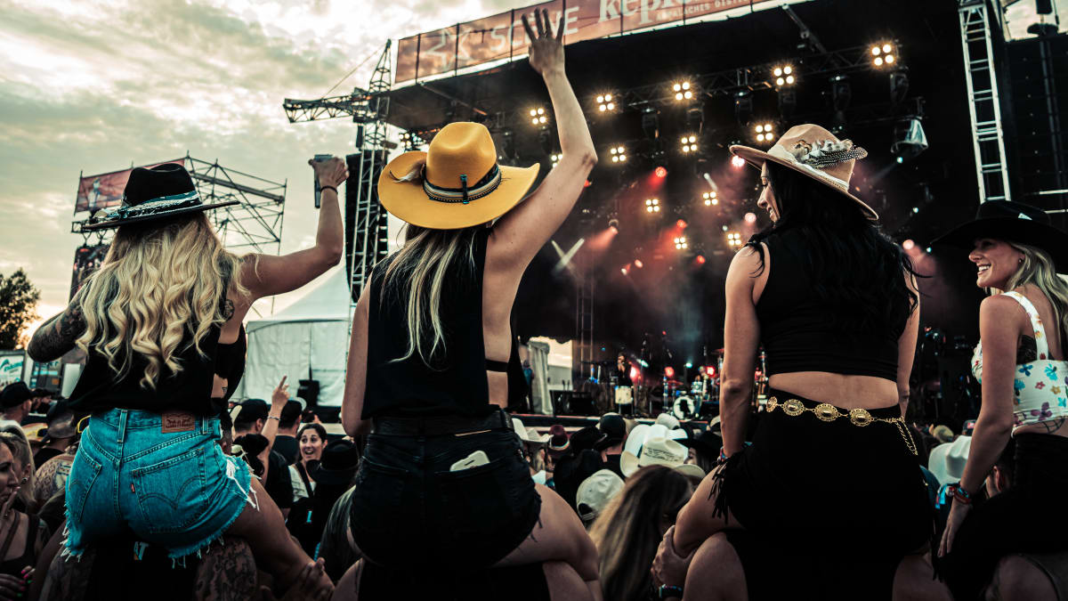 Crowd in front of an outdoor stage including four women wearing cowboy hats.
