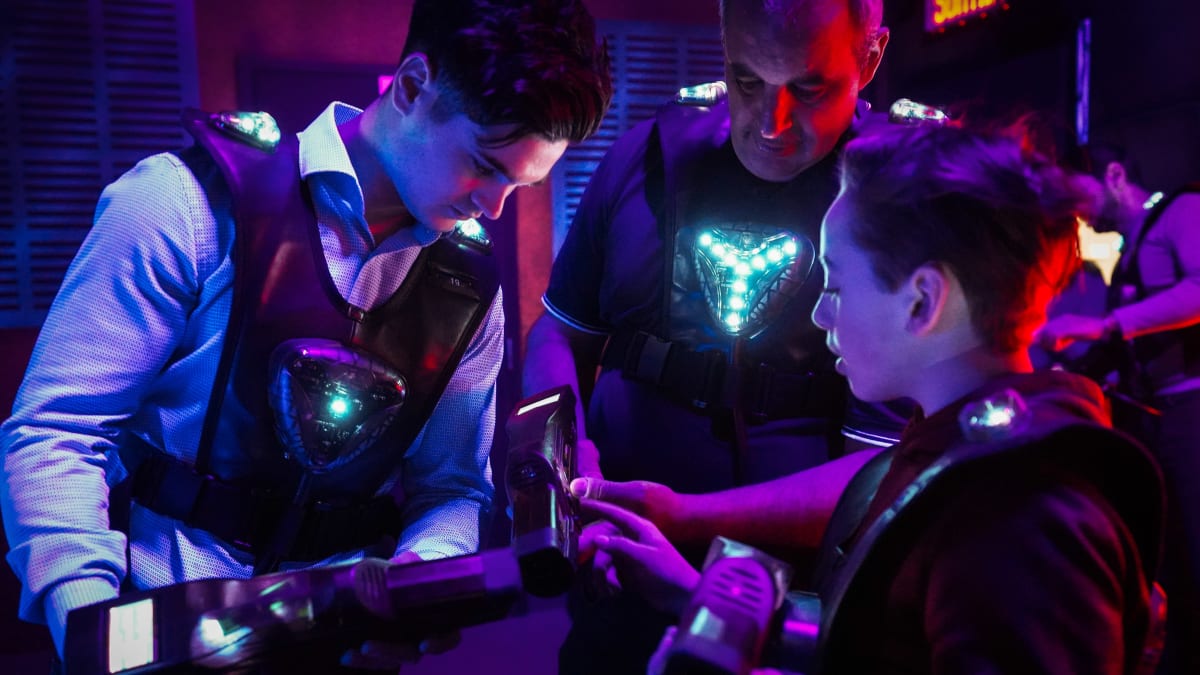 Three people of varying ages grouped together in a laser tag arena, equipped with glowing vests and laser guns.