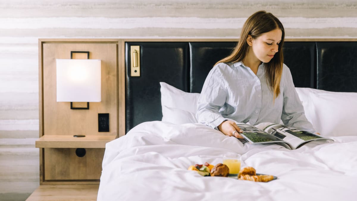 A woman in pyjamas is sitting on a bed. She's reading a magazine and there's a breakfast tray on the comforter.