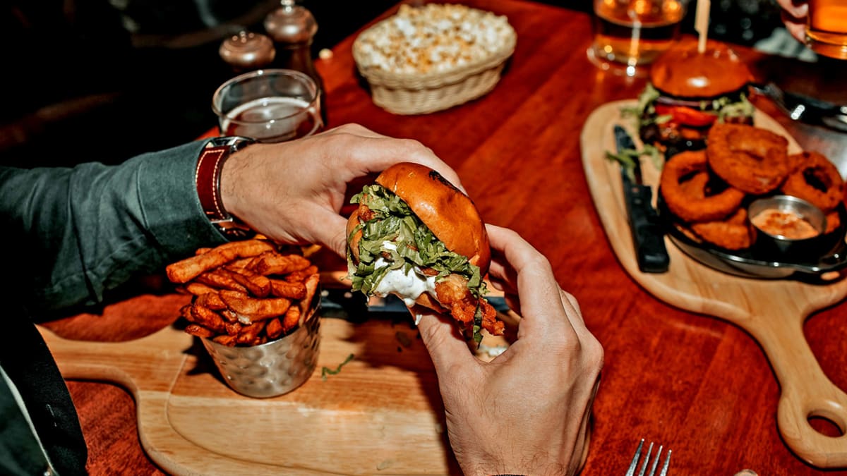 Two hands holding a hamburger above a board with fries on a restaurant table.