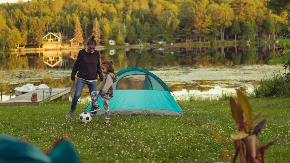 A mother and daughter playing soccer next to a tent by a lake.