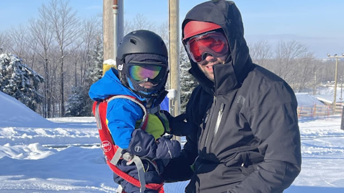 Un père avec son garçon, le bambin a une planche à neige aux pieds et le père est agenouillé devant lui.