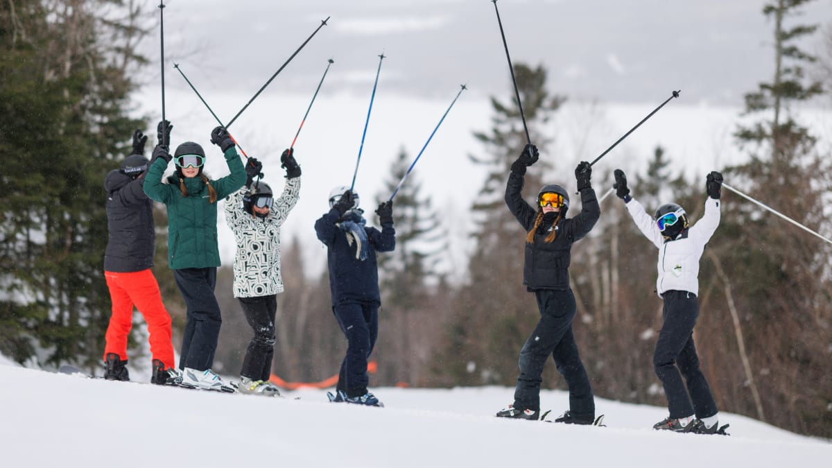 6 skiers at the top of the mountain proudly lifting their poles in the air.