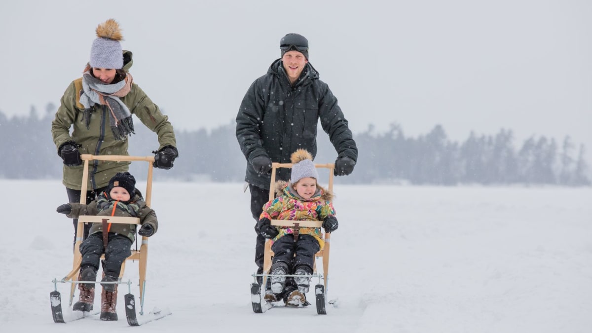 Deux parents qui poussent deux enfants dans des trottinettes sur neige.