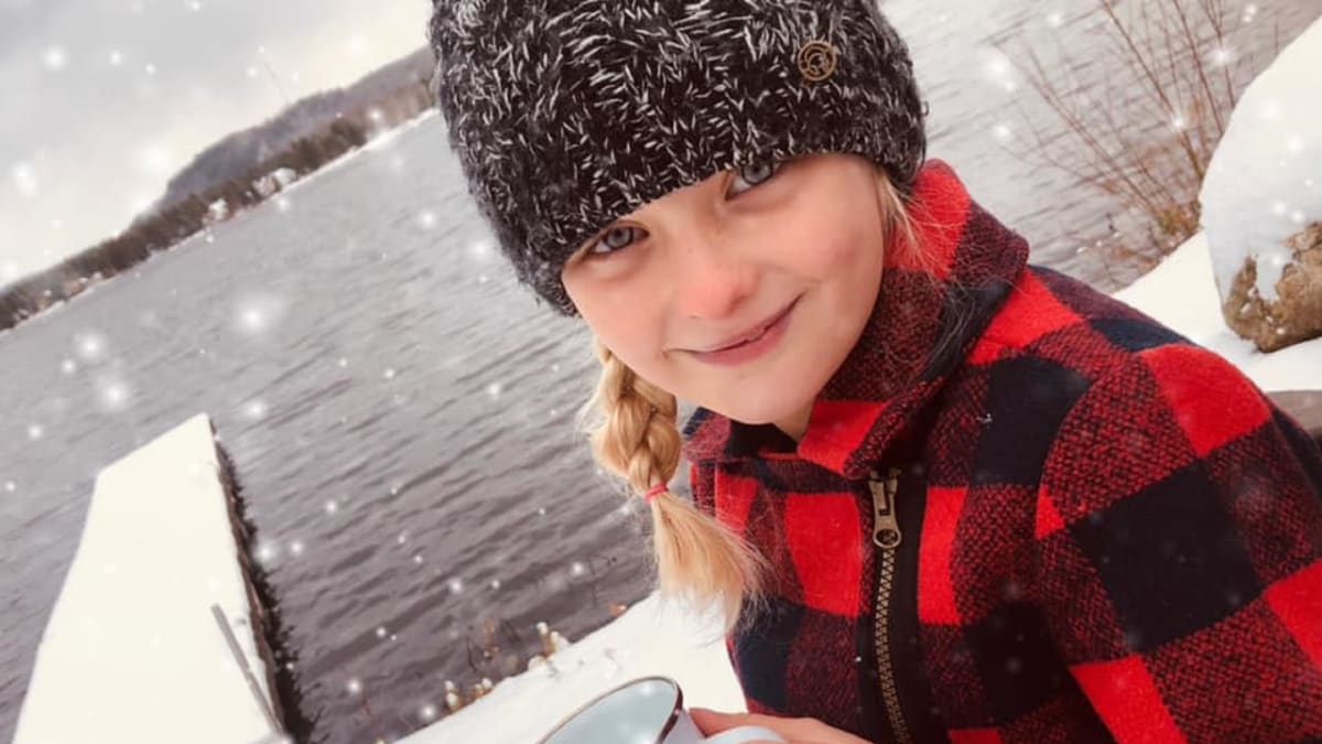 Young girl with a hat and plaid shirt by the water in the winter holding a mug