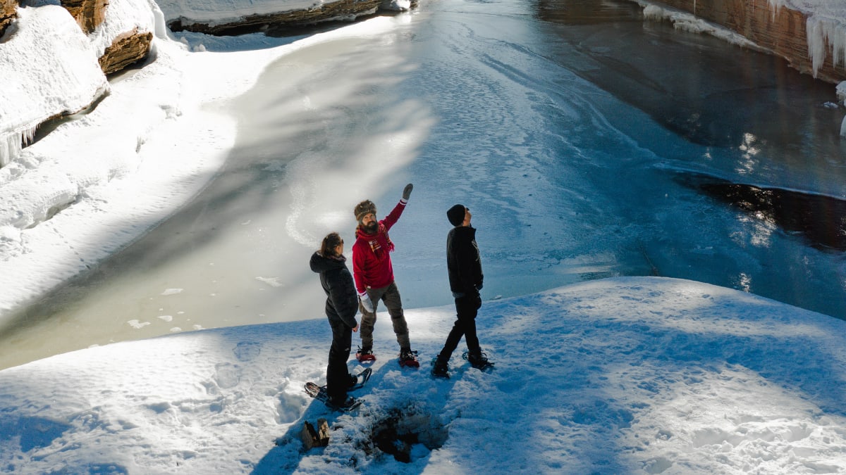 3 people wearing snowshoes crossing a river in the forest in winter.
