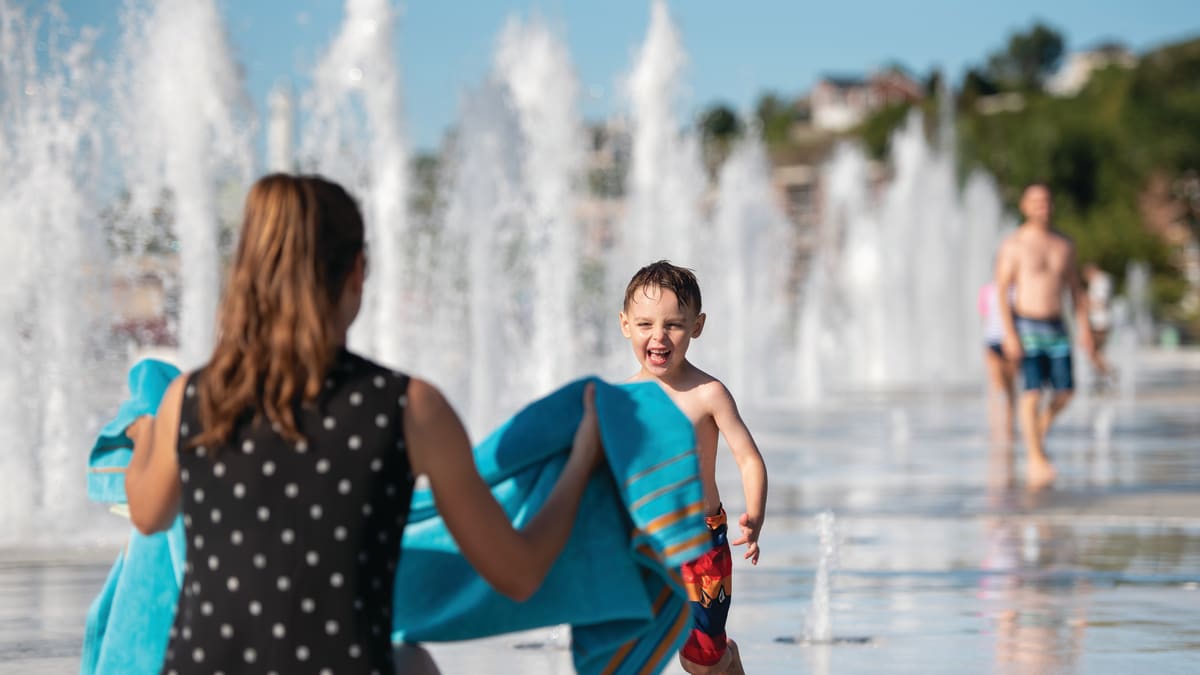 Young boy in water playground happily running towards his mother, who is holding a towel.