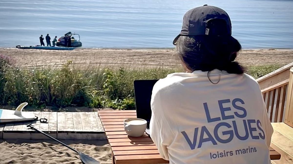 Woman wearing a Les Vagues T-shirt sitting at a café table by the water.