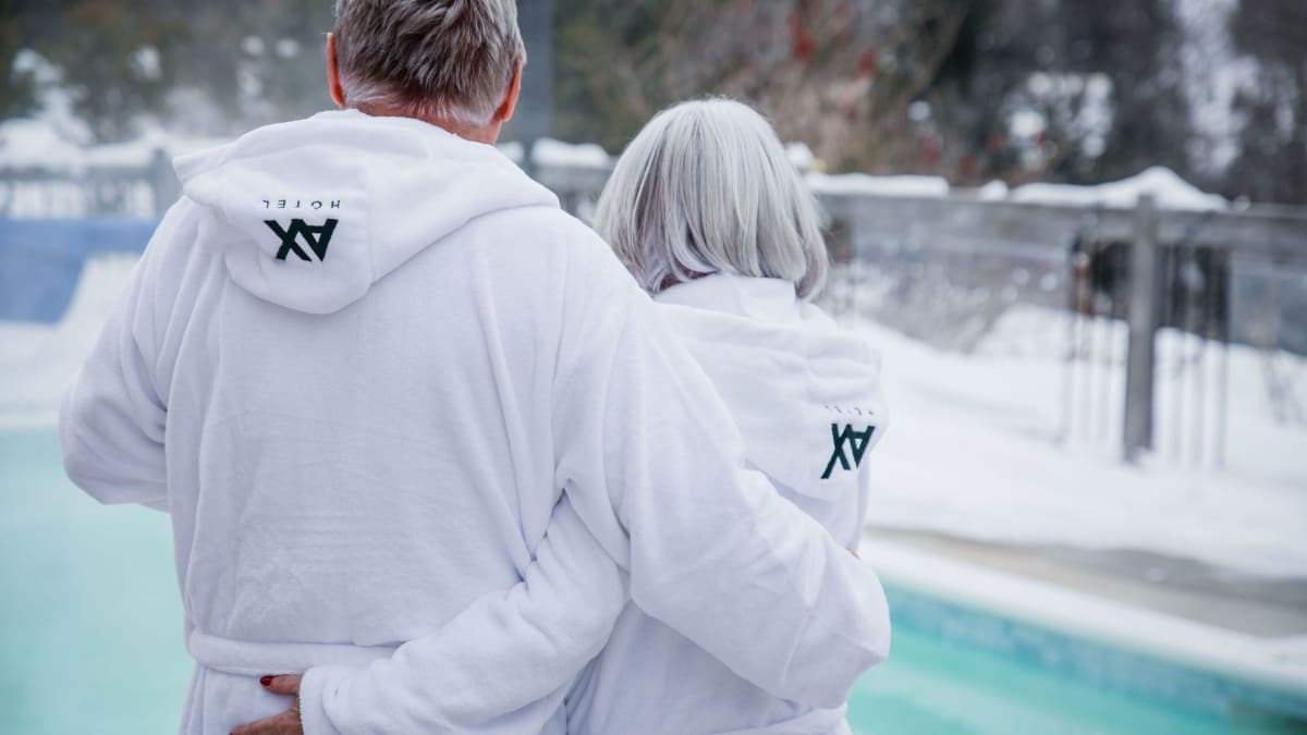 A couple in bathrobes embracing in front of a heated pool surrounded by snow.