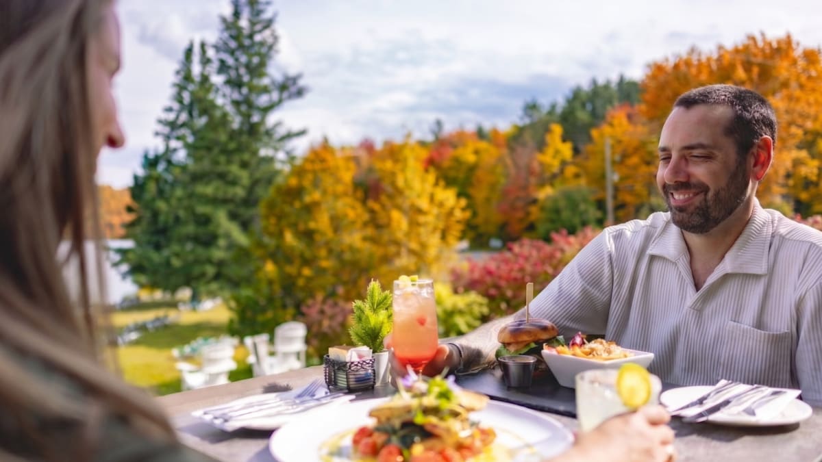 A couple sitting at a table on an outdoor terrace at the Grand Lodge Mont-Tremblant; they are surrounded by a landscape bathed in fall colours.