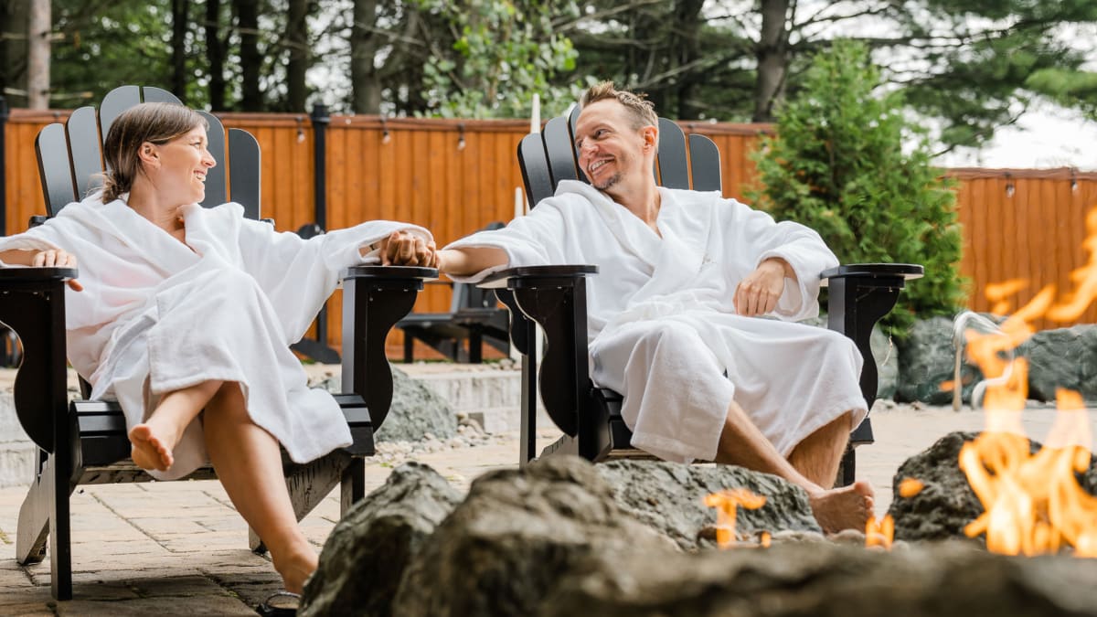 A man and a woman wearing bathrobes, sitting outside on Adirondack chairs by a fire.