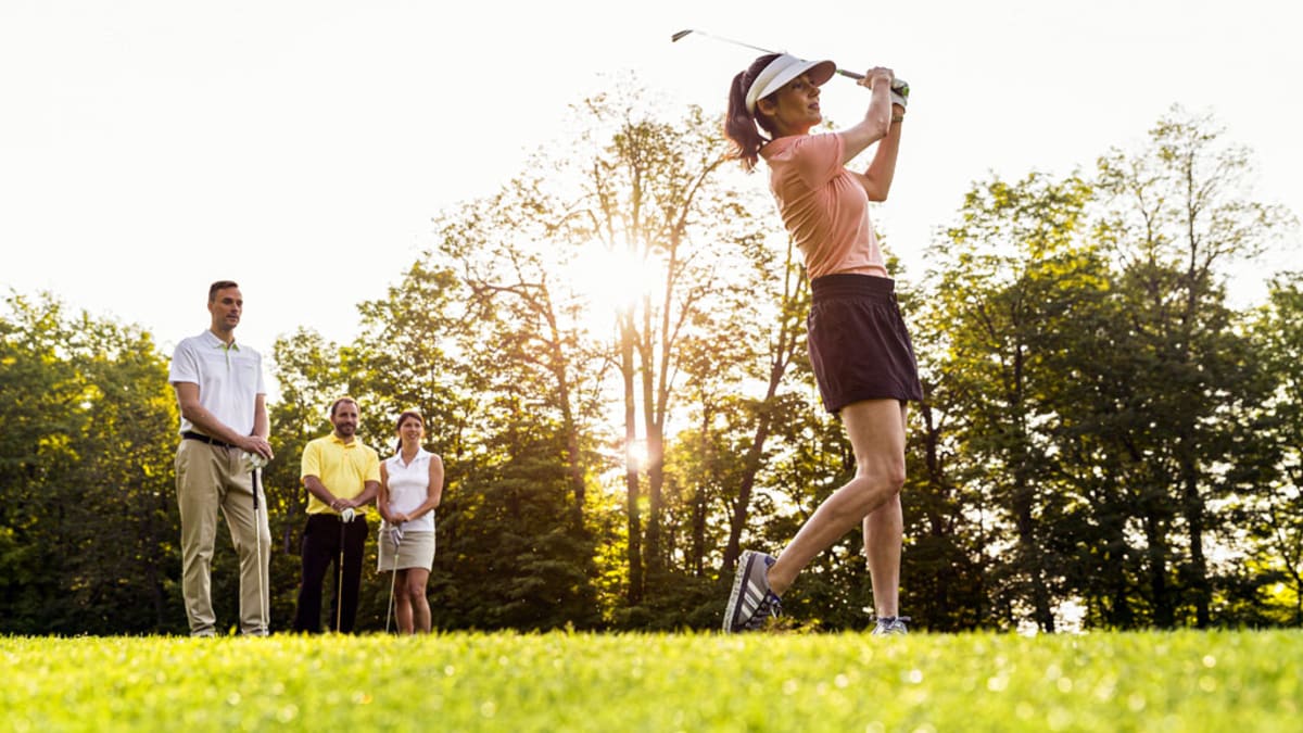 A woman plays golf, with three other individuals observing her in the background.