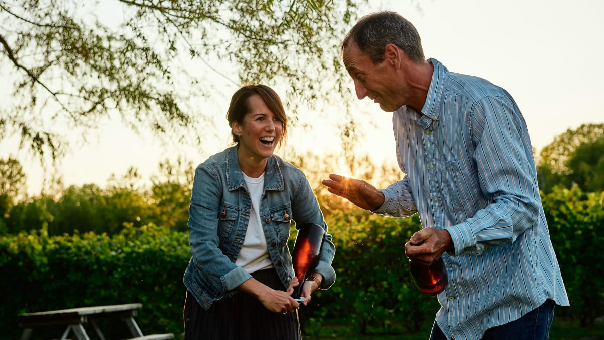 At sunset, a smiling woman and man are each holding a bottle of wine.