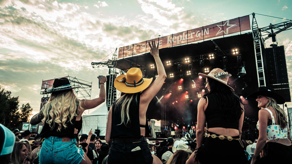 Crowd in front of an outdoor stage including four women wearing cowboy hats.
