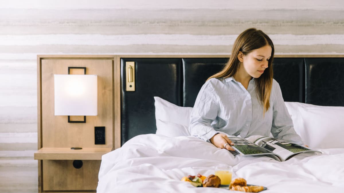 A woman in pyjamas is sitting on a bed. She's reading a magazine and there's a breakfast tray on the comforter.