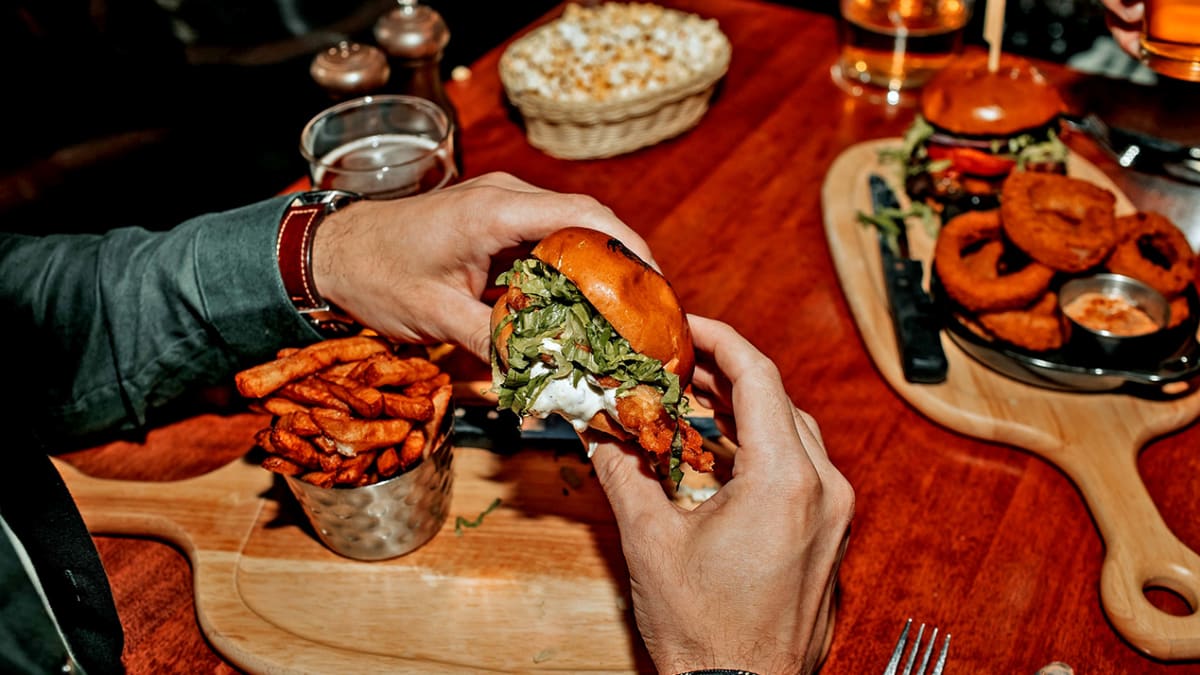 Two hands holding a hamburger above a board with fries on a restaurant table.