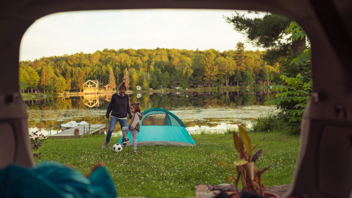 A mother and daughter playing soccer next to a tent by a lake.