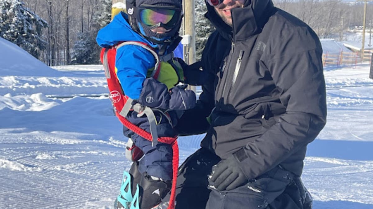 A father with his son, the toddler has a snowboard on his feet and the father is kneeling in front of him.