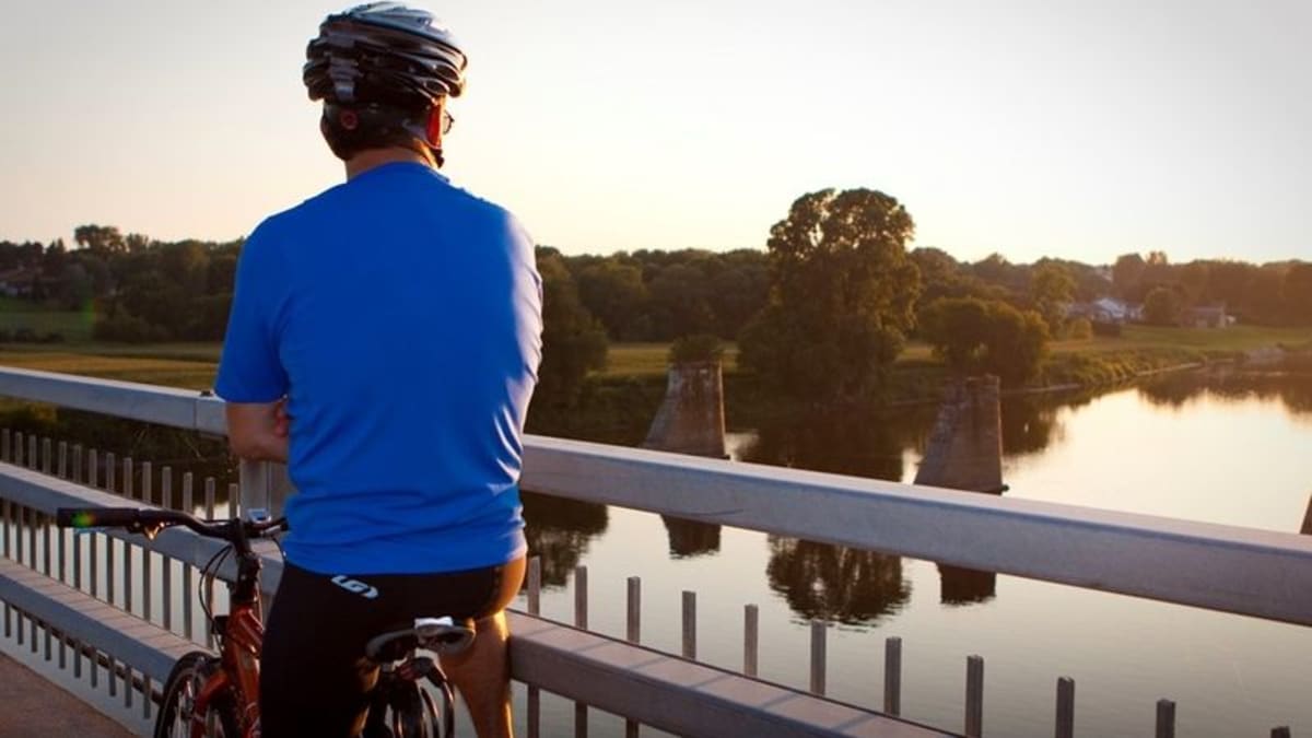 Homme à vélo vu de dos; il semble de tenir sur un pont au-dessus de l’eau.